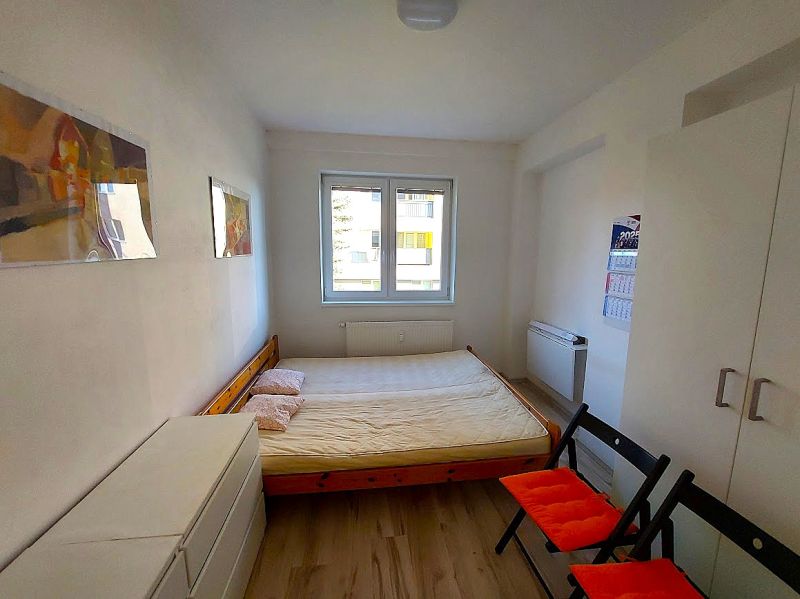 Studio apartment with a bed, white wardrobe, wood-patterned flooring, two folding chairs.