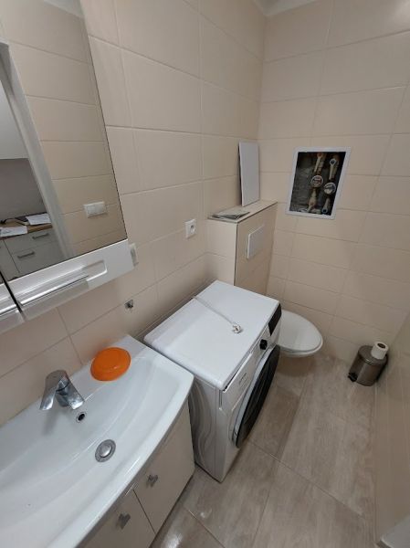 A bathroom in a studio apartment with a washing machine, sink, and toilet, light wall tiles.
