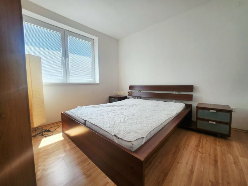 A bedroom with sunlight, a bed, a nightstand, and a wooden-patterned floor.