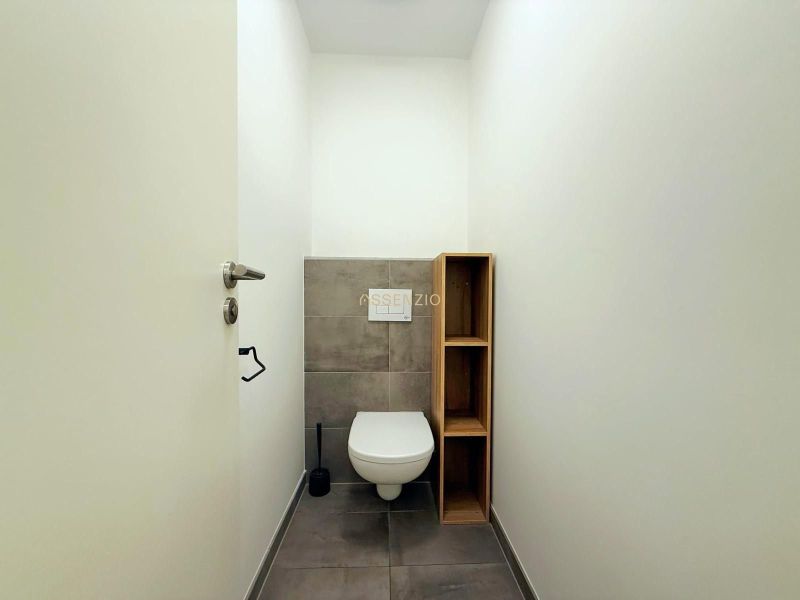 A toilet with gray tiles and a wooden shelf in a two-room apartment.