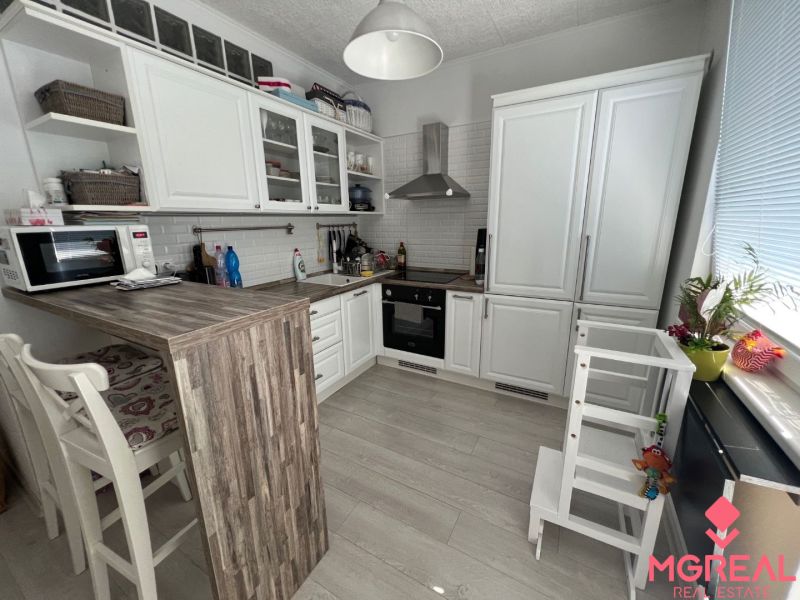 A kitchen in a 2-room apartment with white furniture, a bar counter, and a wooden decor floor.