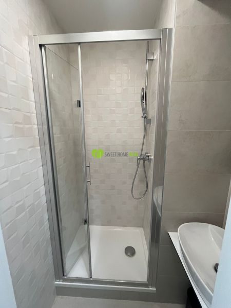 A shower corner and a sink in the bathroom of a studio apartment with simple white tiles.