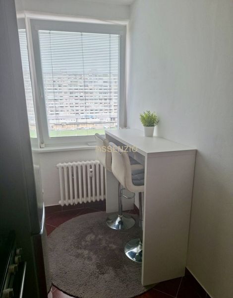 A kitchen in a 3-room apartment with white furniture, a window with blinds, and a flower.
