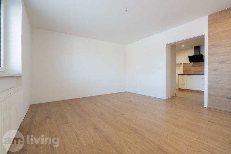 A room in a family house with white walls, a wooden decor floor, and a kitchen.