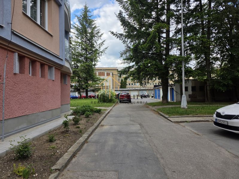 The exterior of an apartment building in Banská Bystrica with an adjacent parking lot and greenery.