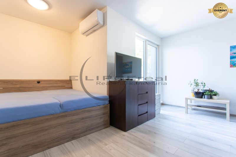 A bedroom in a family house with a bed, TV, and plants on a light floor with a wooden decor.