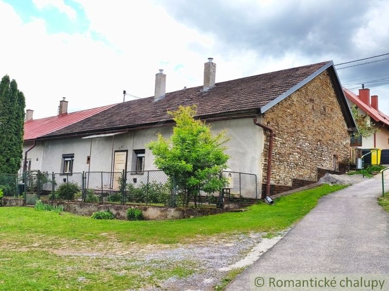 A cottage in Hermanovce with a stone wall and a garden by an asphalt road.