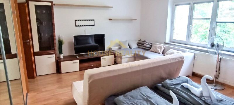 A one-room apartment with a sofa, television, large window, and wooden decor flooring.