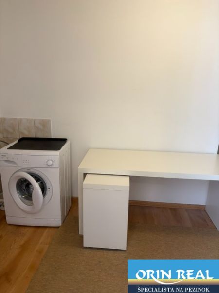 A spacious room in a one-bedroom apartment with a washing machine and a white table, wooden decor flooring.