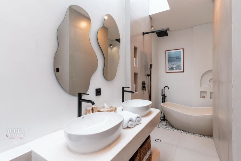 A modern villa bathroom with a marble bathtub, sinks, and mirrors of irregular shapes.