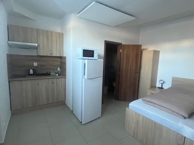 The interior of a building with a kitchenette, refrigerator, microwave, and bed in light shades.