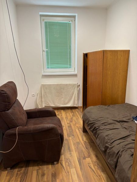 A room with a window, a brown armchair, a bed, and a wardrobe, with a wooden decor floor.