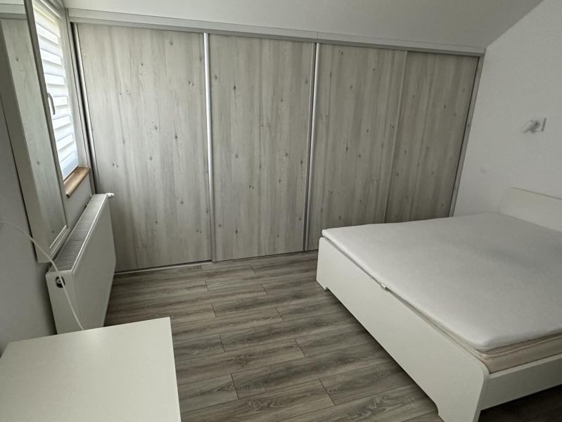 A bedroom in a one-room apartment with a white bed, built-in wardrobe, and wood-patterned flooring.