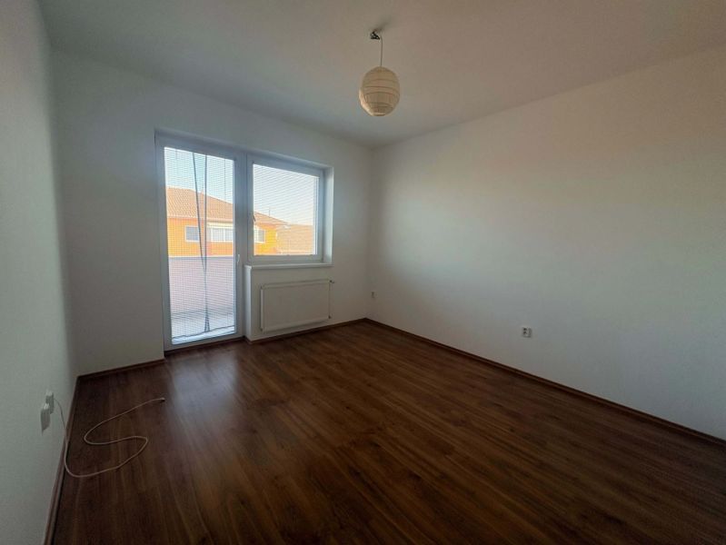 A room in a three-room apartment with a wood-patterned floor and a large window.