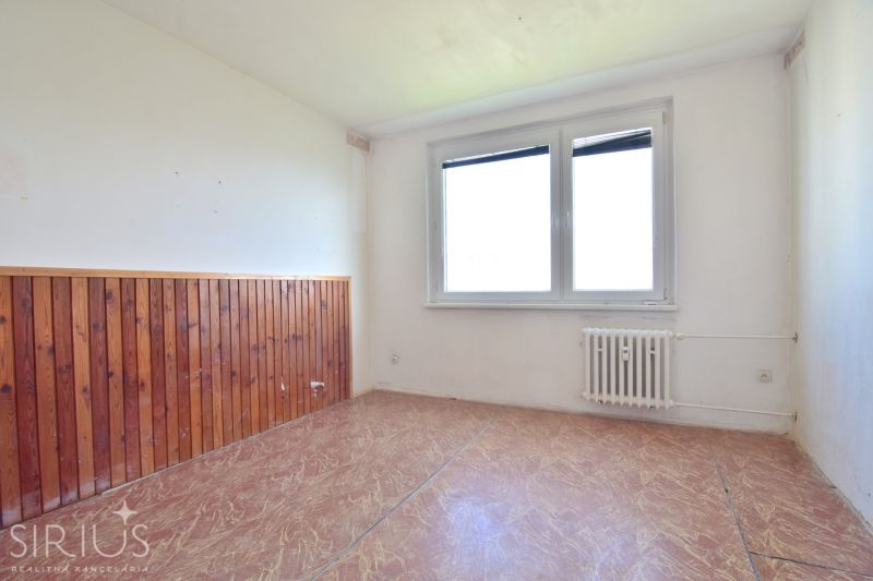 An unfurnished room with a window, a radiator, and a wall with wood paneling in a 3-room apartment.