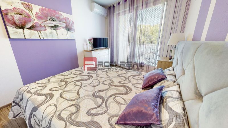 A double bed with purple accessories and a TV in a 2-room apartment with a wood-patterned floor.