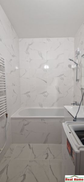 A bathroom in a 2-room apartment with a bathtub and a washing machine, tiled with light tiles.