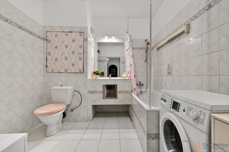 Bathroom in a 2-room apartment with a washing machine, bathtub, toilet, and sink with a floral cabinet.