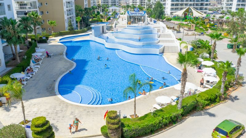 A pool in Iskele surrounded by palm trees and loungers near apartment buildings.