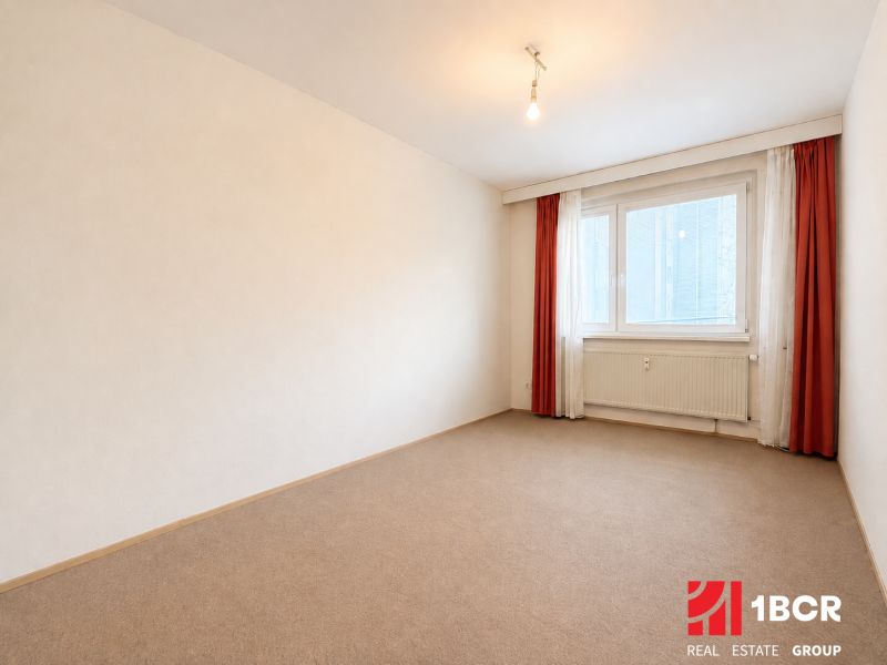 A room with carpet flooring and a window in a 3-room apartment, simple curtains.