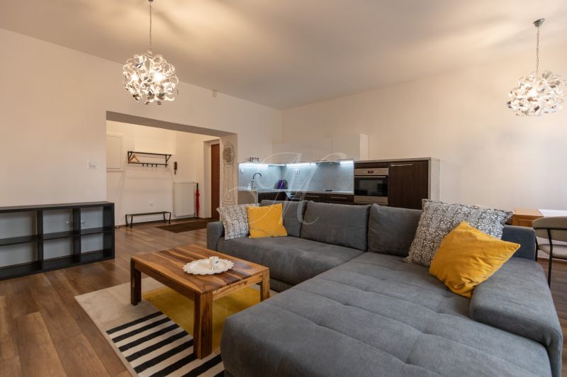 Living room with a kitchen, gray sofa, wooden table, and floor with a wooden design.