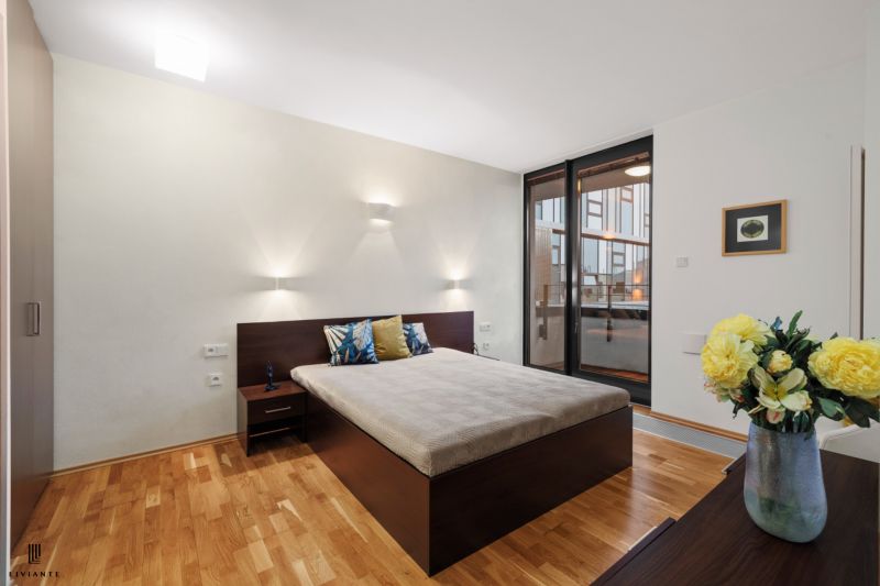Bedroom in a 2-room apartment with a bed, wood-patterned flooring, and large windows.