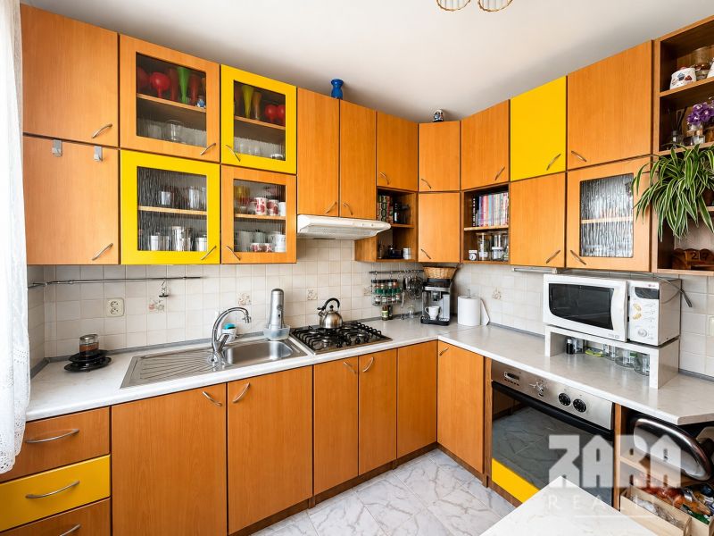 A kitchen in a 3-room apartment with a wooden decor and yellow cabinets, appliances, and a plant.