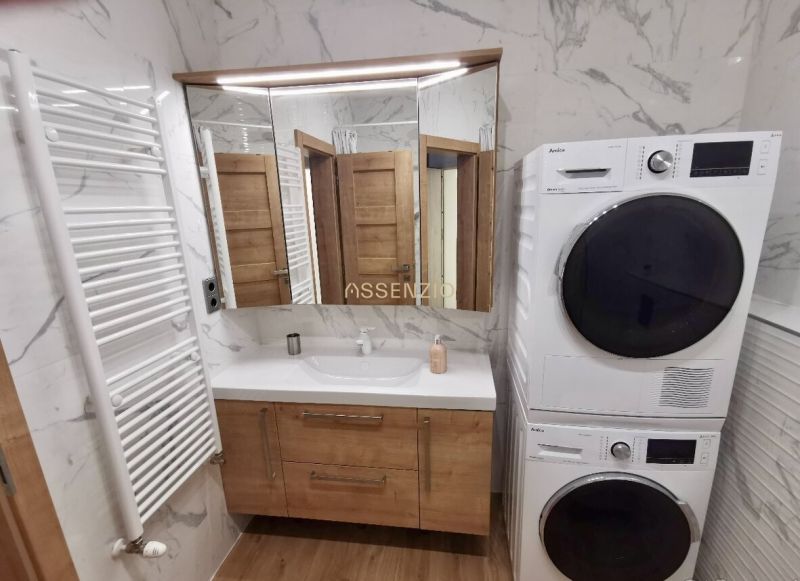 A bathroom in a 2-room apartment with a sink, washing machine, dryer, and a floor with a wooden decor.
