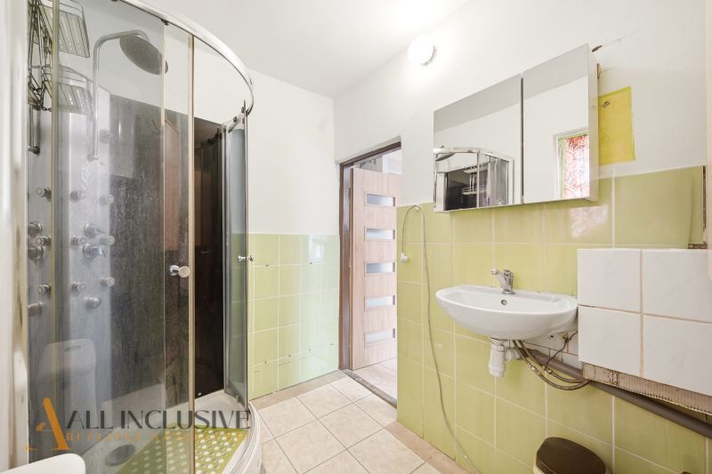 A bathroom with a corner shower, green tiles, a sink, and a mirror in a family house.