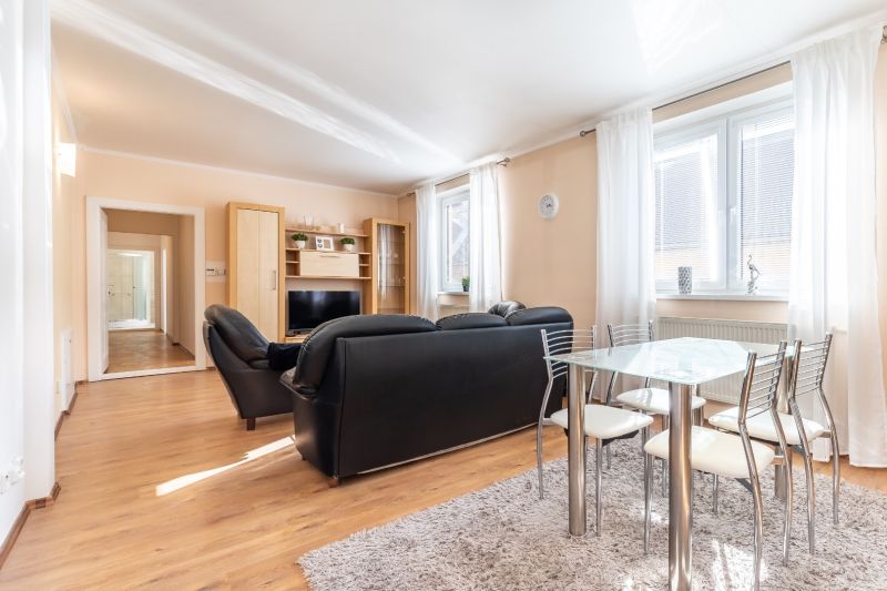 Living room in a 3-room apartment with a black sofa, glass table, and wooden decor flooring.