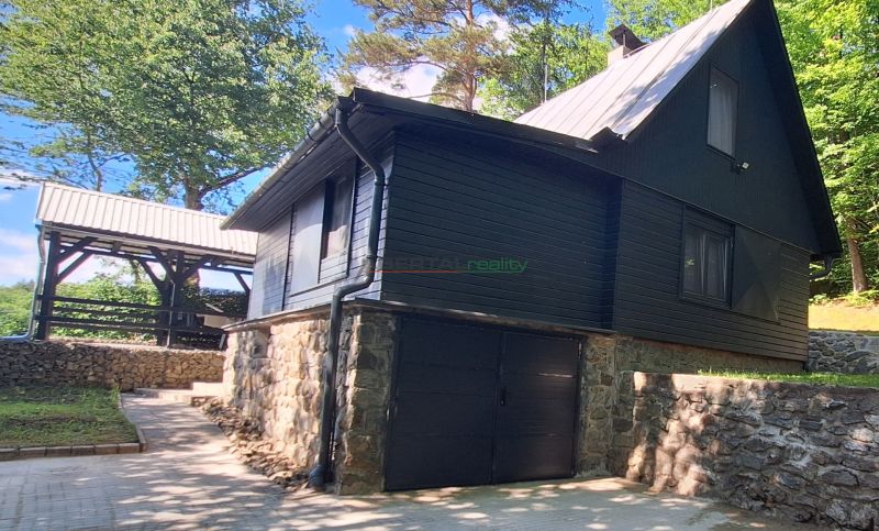 Black cottage in the village of Bukovec with a stone foundation, surrounded by greenery and a shelter.