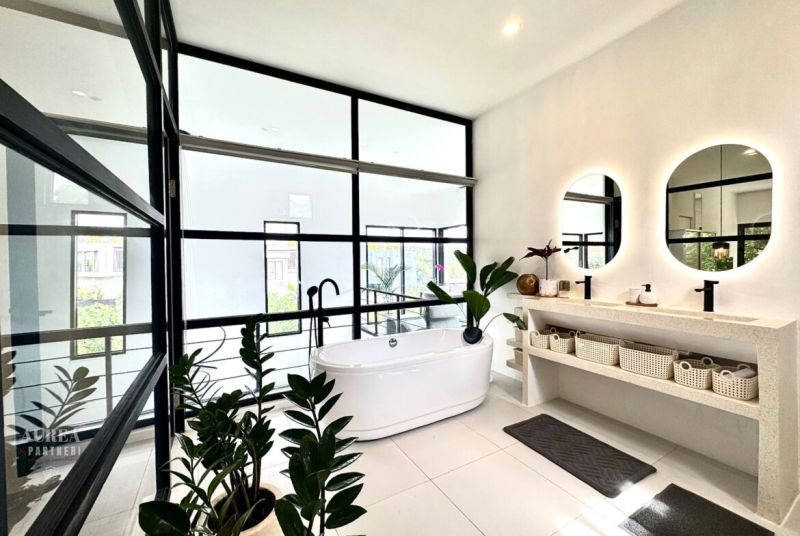 A bathroom in a villa with a bathtub, large windows, round mirrors, and houseplants.