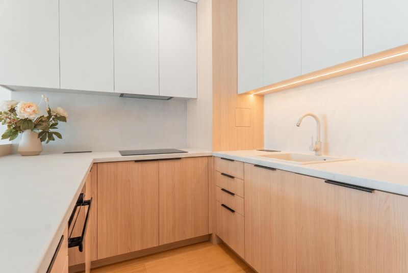 A kitchen in a 3-room apartment with white cabinets and a sink on a floor with a wood decor.