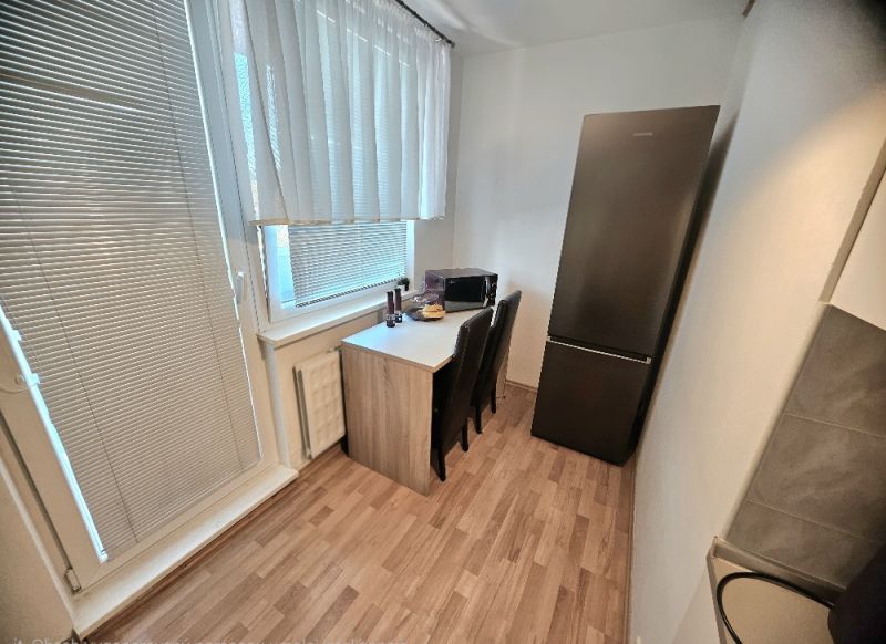 A kitchen in a studio apartment with a wood-patterned floor, a refrigerator, and a table by the window.