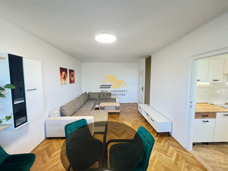 Bright living room with a sofa, wood-patterned flooring, and a kitchen in a 2-room apartment.