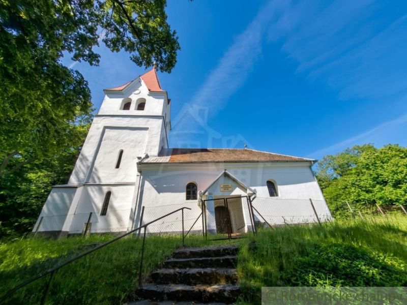 The church in the village of Ozdín with a tower and staircase, surrounded by greenery and a blue sky.