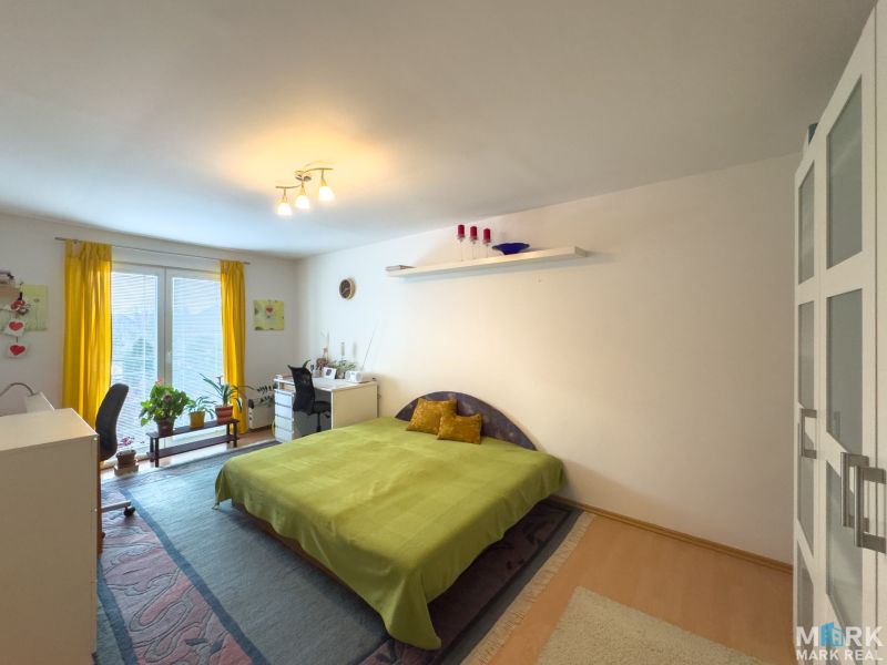 A bright bedroom in a family house with a bed, a desk, and a wooden-patterned floor.
