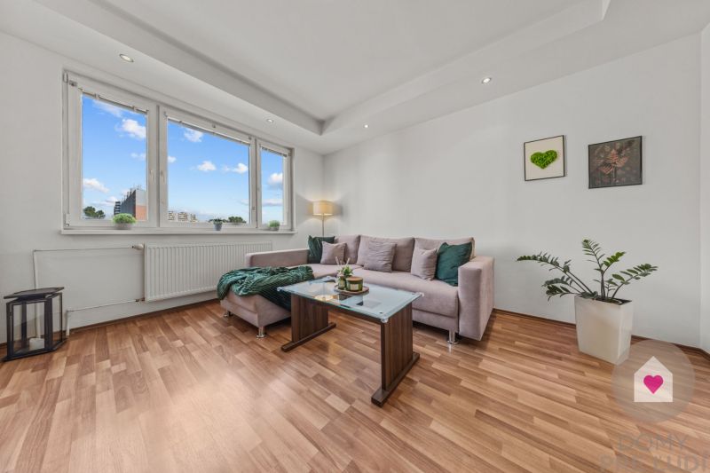 Living room in a 4-room apartment with a sofa, wooden decor flooring, plants, and windows.