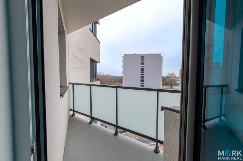 A balcony in a 2-room apartment with a view of the surrounding buildings through a glass railing.