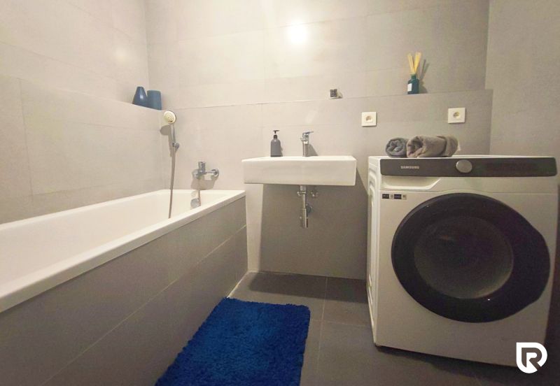 Bathroom in a 2-room apartment with a bathtub, sink, and washing machine on a tiled floor.