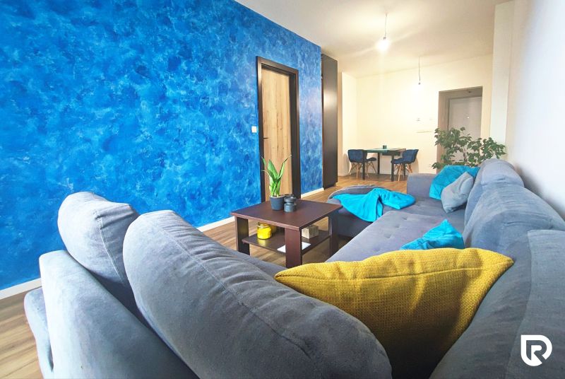 Living room in a two-room apartment, gray sofa, blue wall, wooden floor decor, dining area.