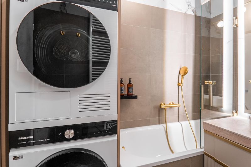 A bathroom in a 3-room apartment with a washing machine, dryer, bathtub, and gold accessories.