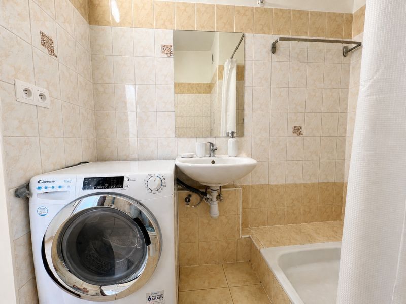 Bathroom in a studio apartment with a washing machine, sink, and shower with a white curtain.