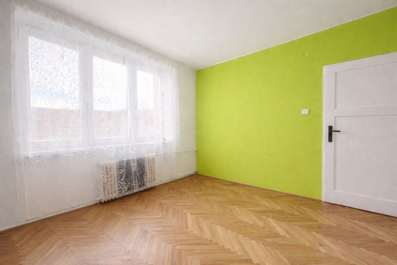 A room with a green wall, white curtains, and a wood-patterned floor in a two-room apartment.