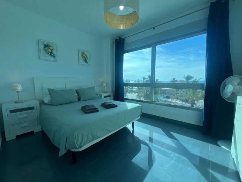 Bedroom of a holiday apartment with a large window, bed, and tables.