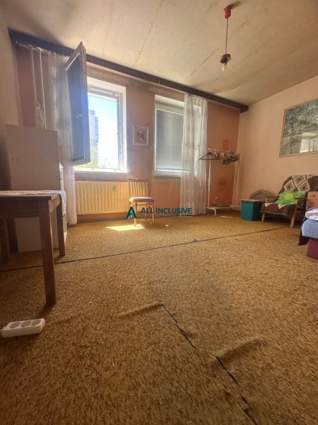 A room in a 2-room apartment with a carpet, an open window, and simple furniture.