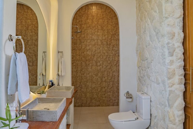 A bathroom in a villa with a stone wall, double sink, and a shower behind an arch.