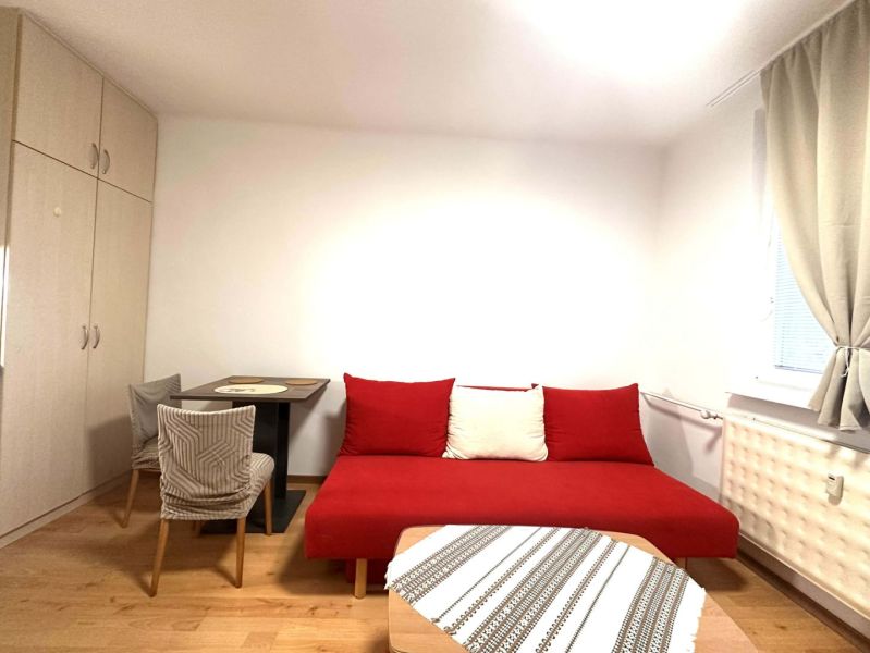 A room in a recreational apartment with a red sofa, a table, and a wooden-patterned floor.
