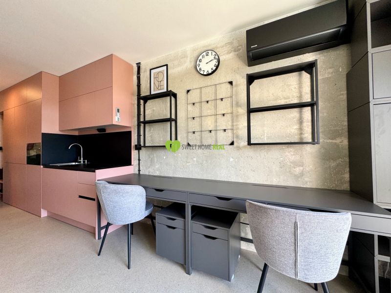 A studio apartment with a pink kitchen and a work desk, flooring with a wood decor.