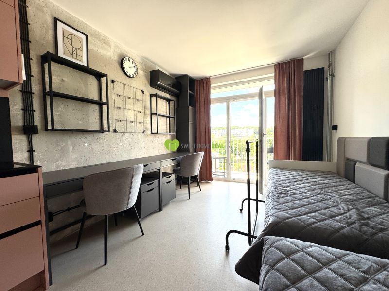 Studio apartment with a desk, bed, shelves, and balcony doors.